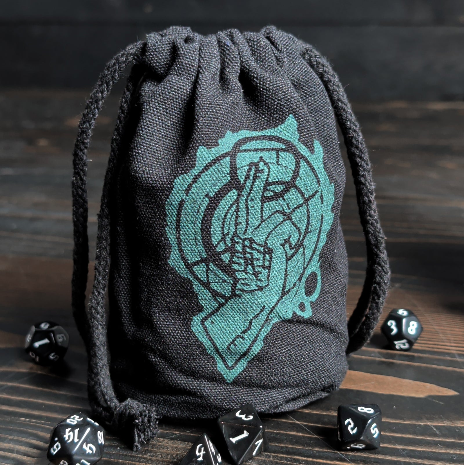 Black drawstring dice bag with an emblem of a hand in the dispel symbol, surrounded by dice on a wooden surface.