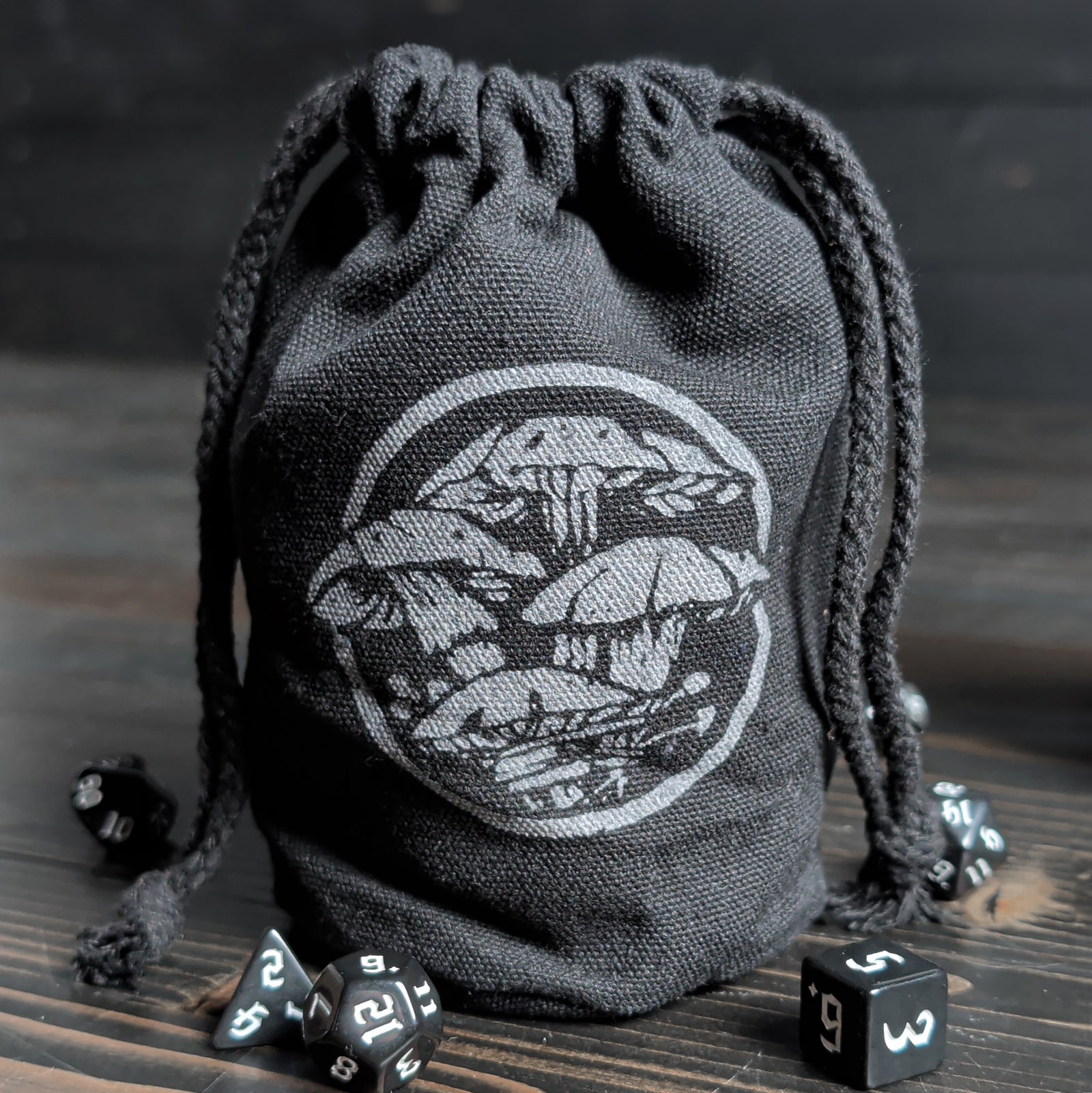 Black drawstring dice bag with a mushroom art on a wooden surface with dice around it