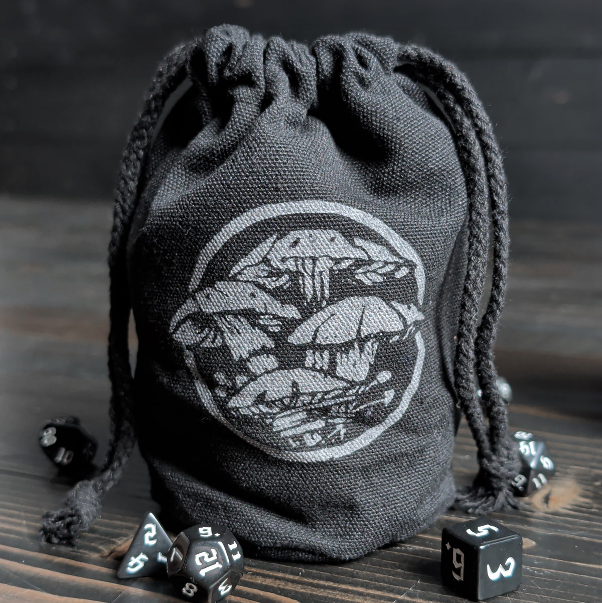 Black drawstring dice bag with a mushroom art on a wooden surface with dice around it