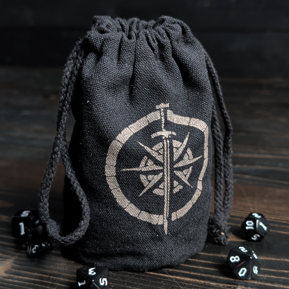 Dark gray drawstring bag with compass design on a wooden surface with dice around it