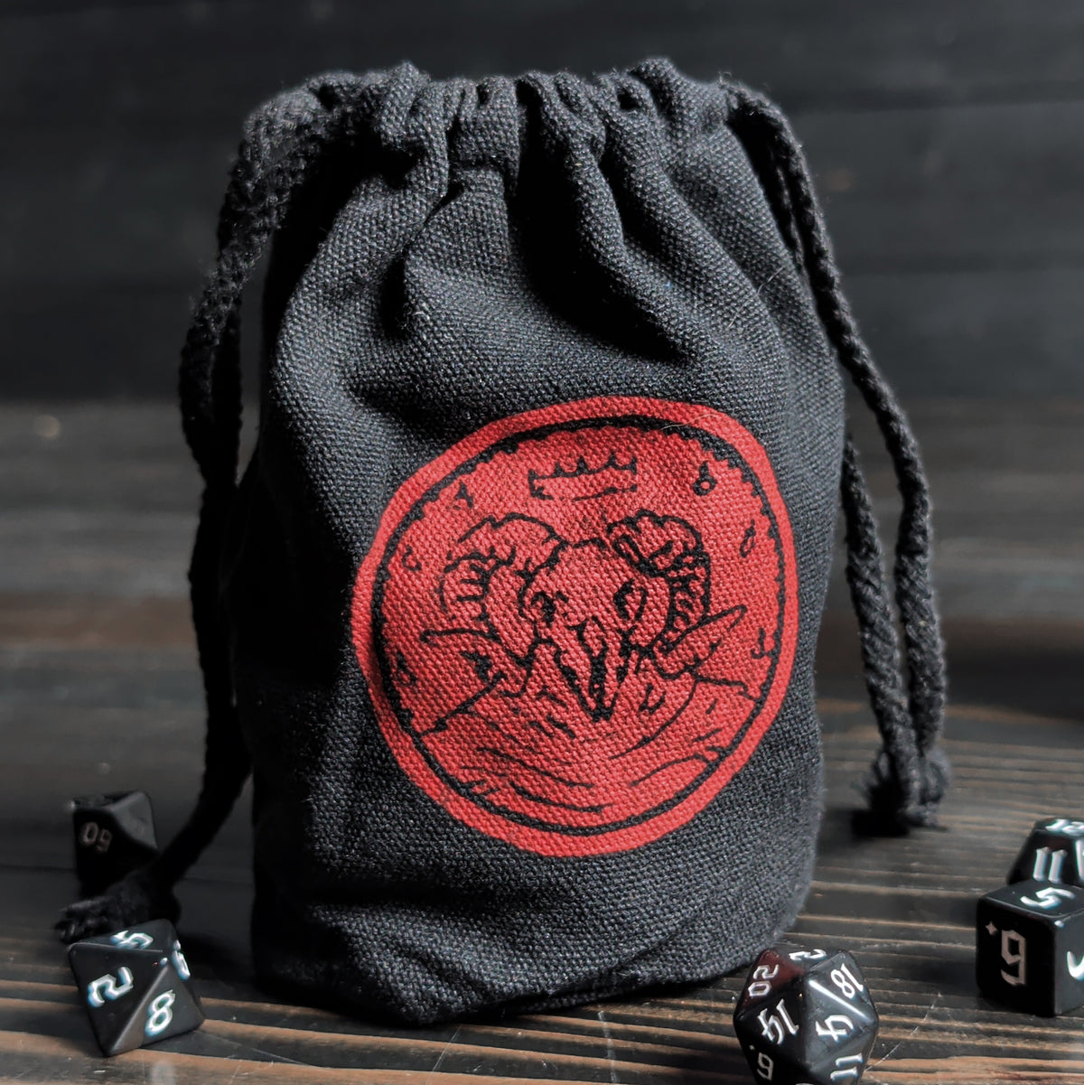 Black drawstring dice bag with red emblem featuring the sigil of the dark one, surrounded by dice on a wooden surface.